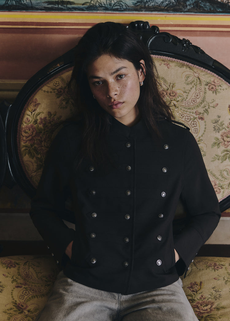 Chaqueta marrón de doble botonadura metálica vista frontal con los puños en los bolsillos